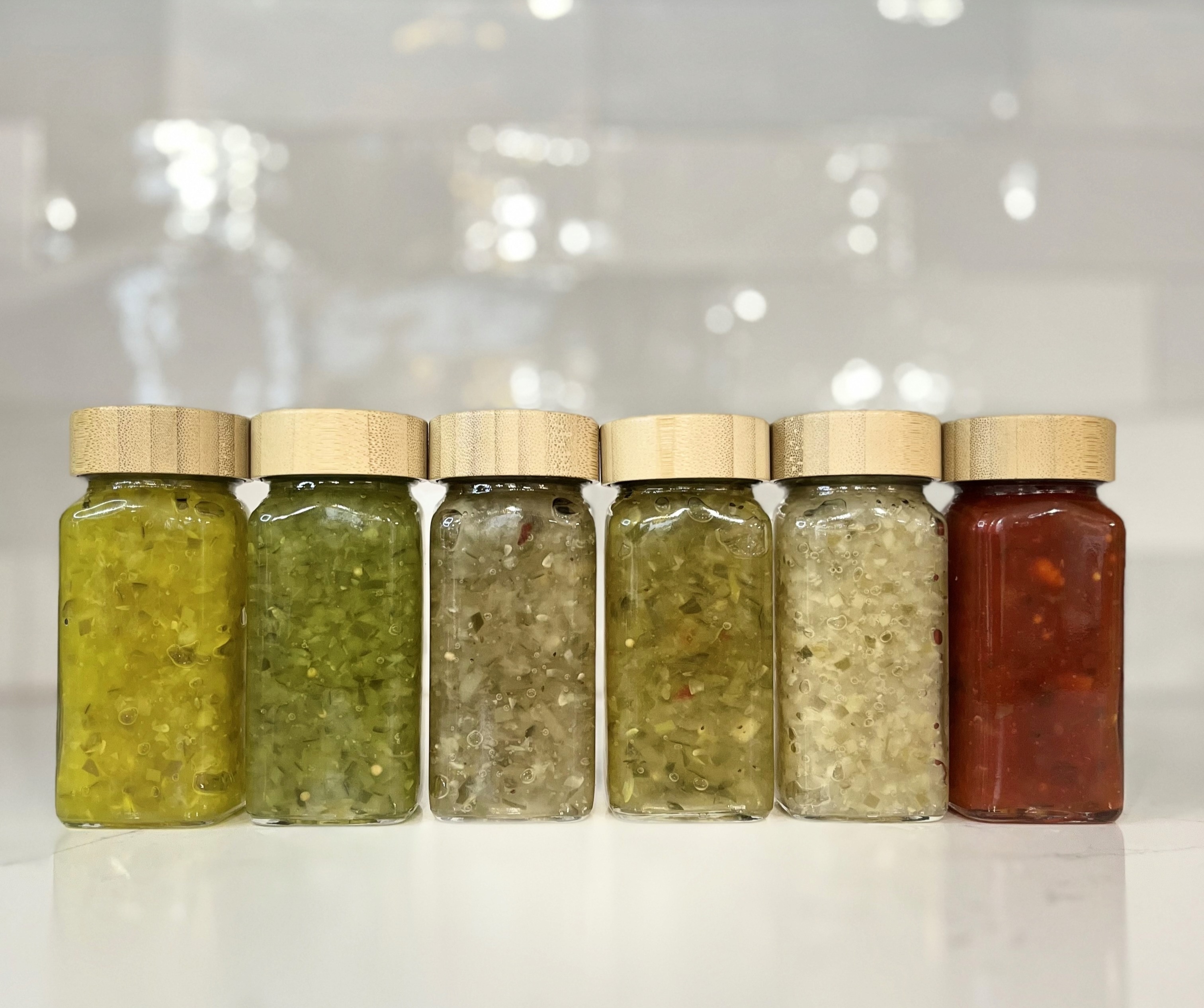 Photo of six glass jars filled with different varieties of Flamm's Pickles relish in shades of green, yellow-green, and red, arranged in a row on a kitchen countertop. 